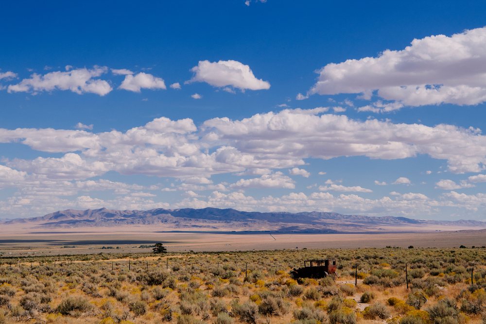 Vast Nevada desert landscape with a rusted vehicle among dry shrubs under blue sky with clouds and distant mountains, USA.