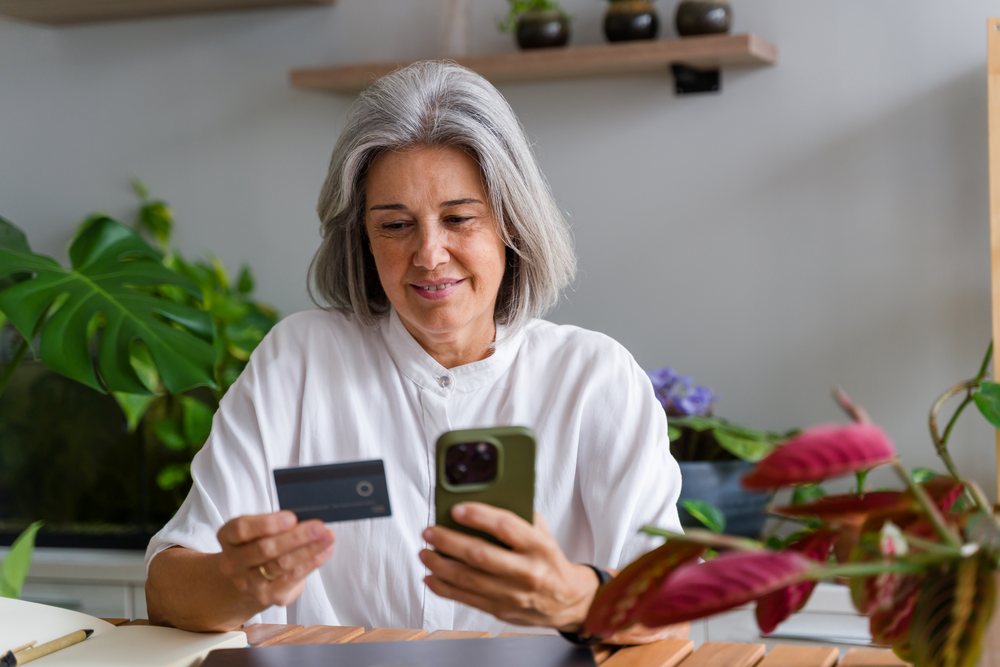 Mature woman with gray hair using a smartphone and holding a credit card at home, surrounded by indoor plants, Spain.