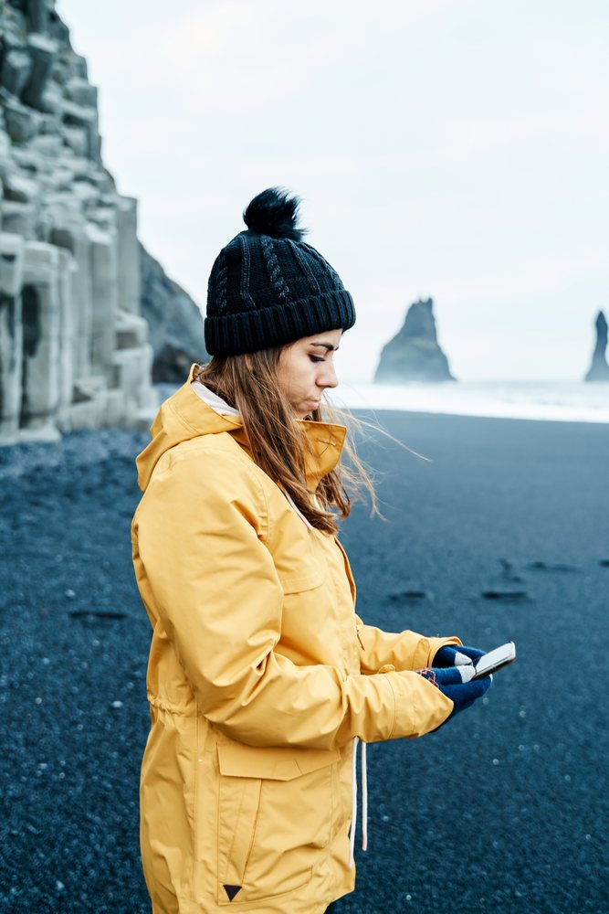 A white woman uses a smartphone on a black sand beach in a beach