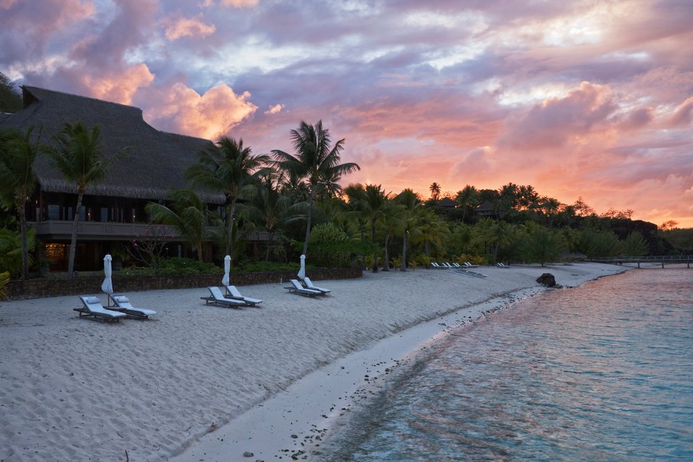Sunrise sky with clouds over a thatched roof Bora Nui Resort beside a tropical sand beach with palm trees, deck chairs, and closed umbrellas, Tahiti, French Polynesia.