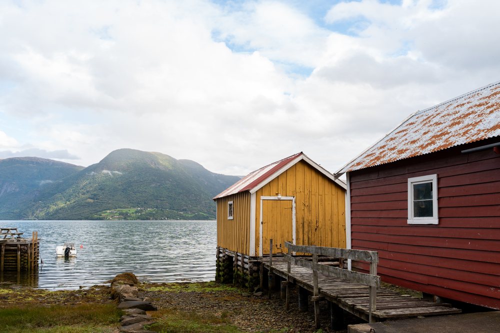 Two wooden boathouses connected by a wooden pier over a rocky shore at Luster Fjord, with mountain landscape and calm water, coastal town, village, Norway.