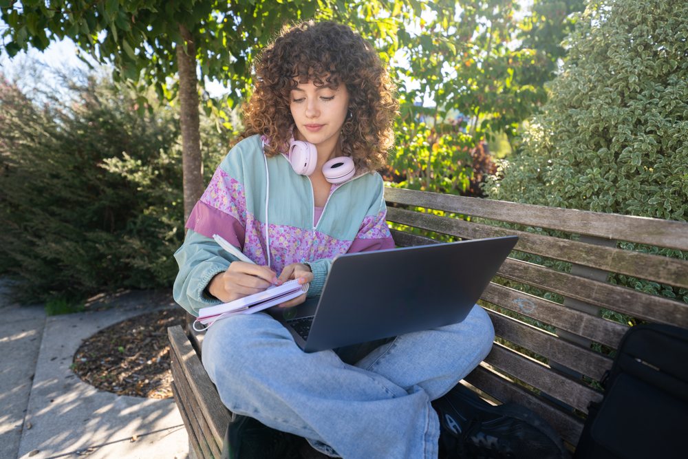 Woman writing in a notebook while seated on a wooden bench in a sunny park with a laptop on her lap, Spain.