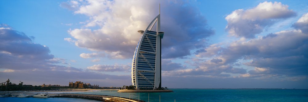 Burj Al Arab hotel stands on its artificial island off Dubai's coast under a partly cloudy sky above the sea and marina.