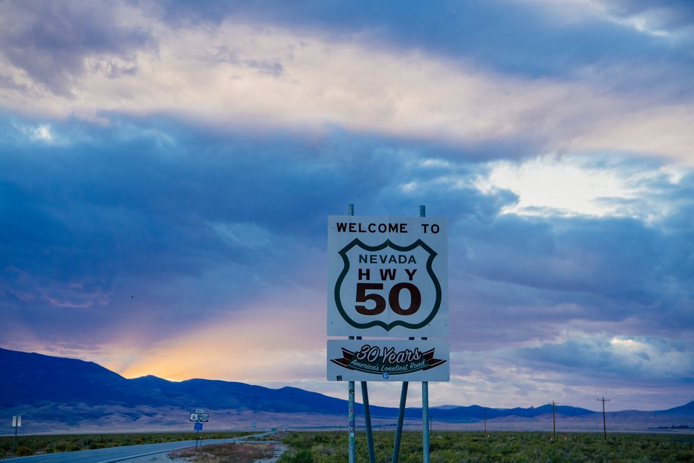 Nevada Highway 50 welcome sign at sunset on an open paved road with mountains and cloudy sky in the background, USA.
