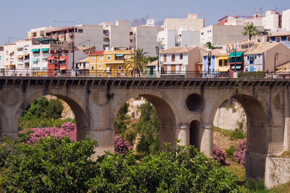 Bridge with arched supports and colorful buildings, with trees and pink flowering plants below on a sunny day, Alicante, Spain.