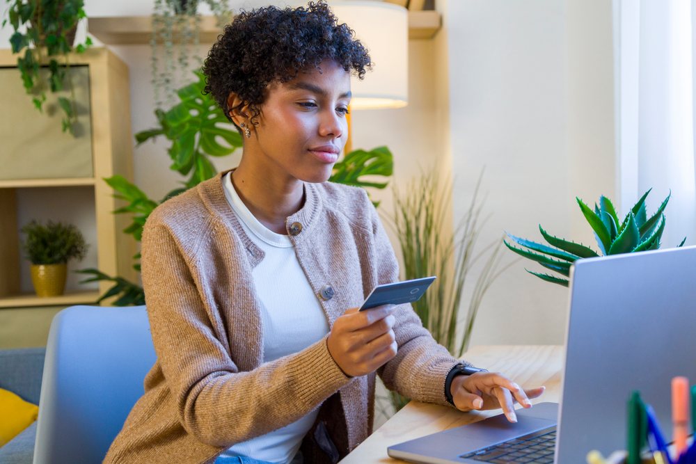 Woman seated at desk in a home office holding a credit card and using a laptop for remote work.