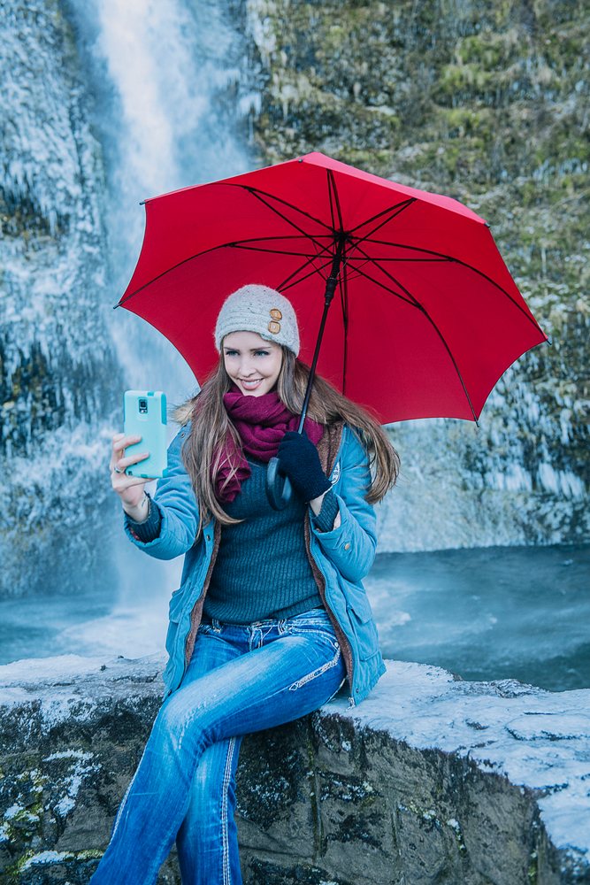 Smiling woman wearing winter clothing sits on a rock wall holding a red umbrella and taking a selfie, Multnomah Falls, Bridal Veil, Oregon.