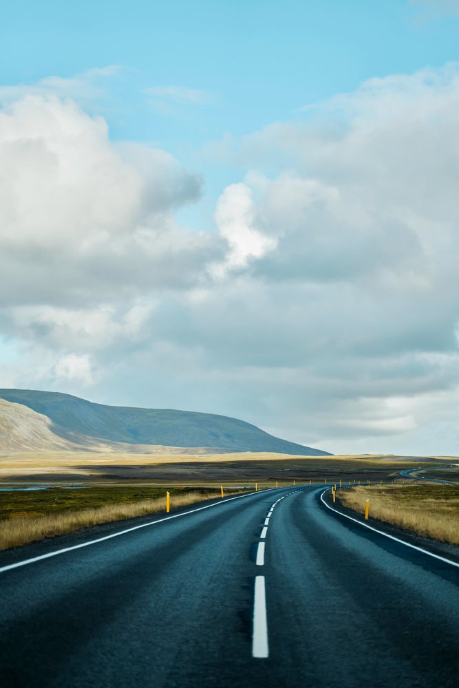 A winding empty asphalt road stretches through open grassland under a blue sky with clouds, Iceland.