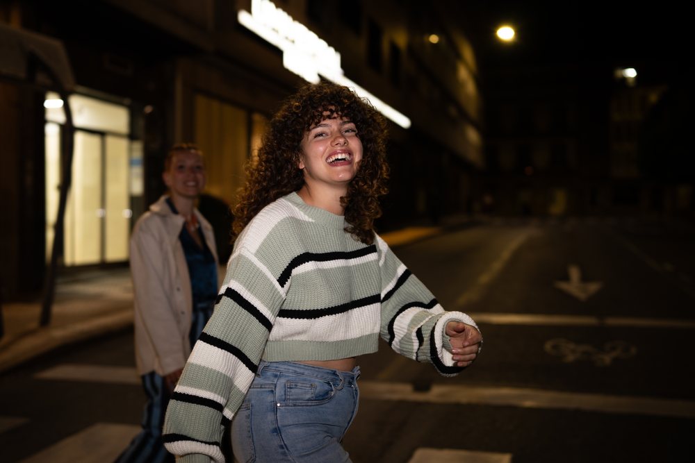 Woman with curly hair laughing while walking on a nighttime city street, accompanied by a friend, friendship, Santiago De Compostela, Spain.