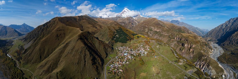 Mountain valley around Gergeti village featuring Kazbegi Peak and snow-capped mountains under a partly cloudy sky, Cityscape, Georgia.