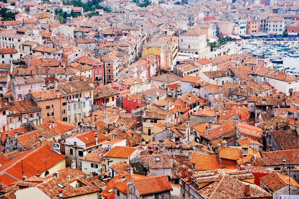 Aerial overview of Rovinj town showing dense red tiled rooftops and boats in the harbor along the Adriatic coast.