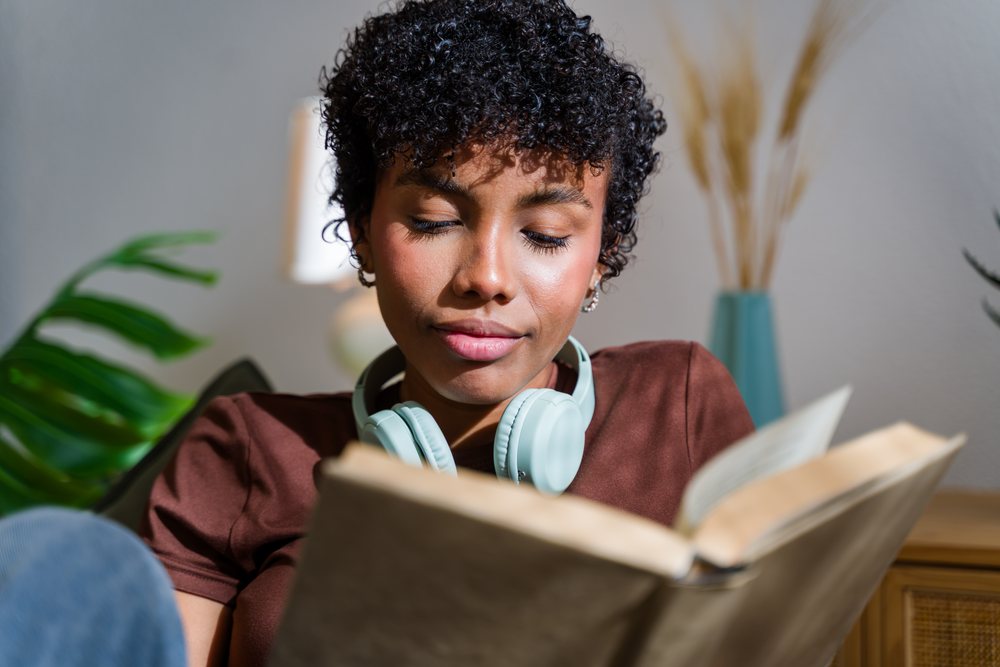 Woman wearing headphones reading a book indoors in a cozy home setting with decorative plants and vases, Spain.