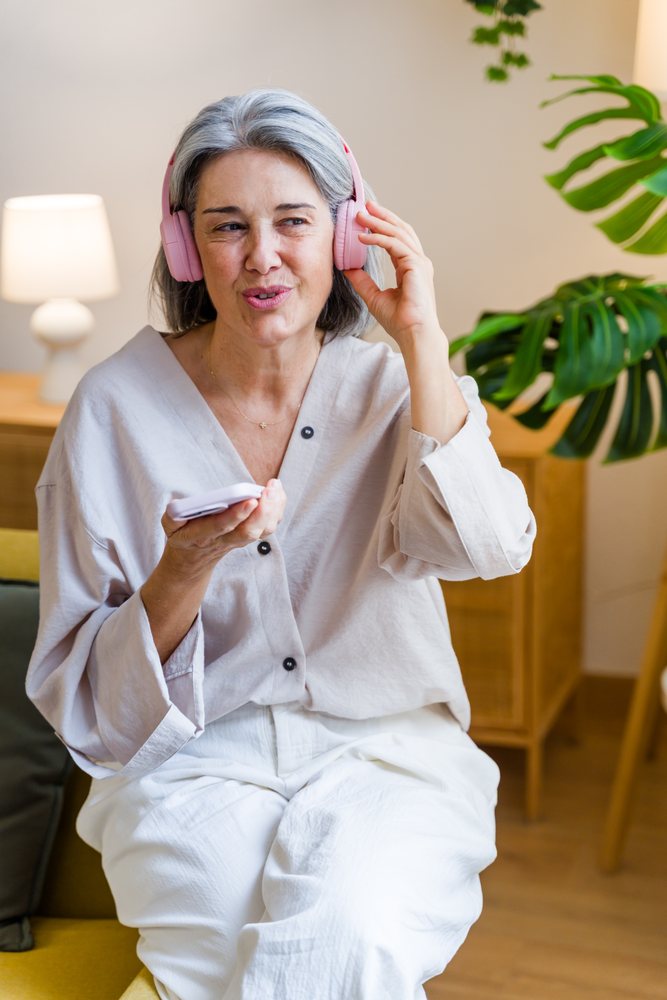 Mature woman with pink headphones listens to smartphone indoors in a modern living room, Spain.