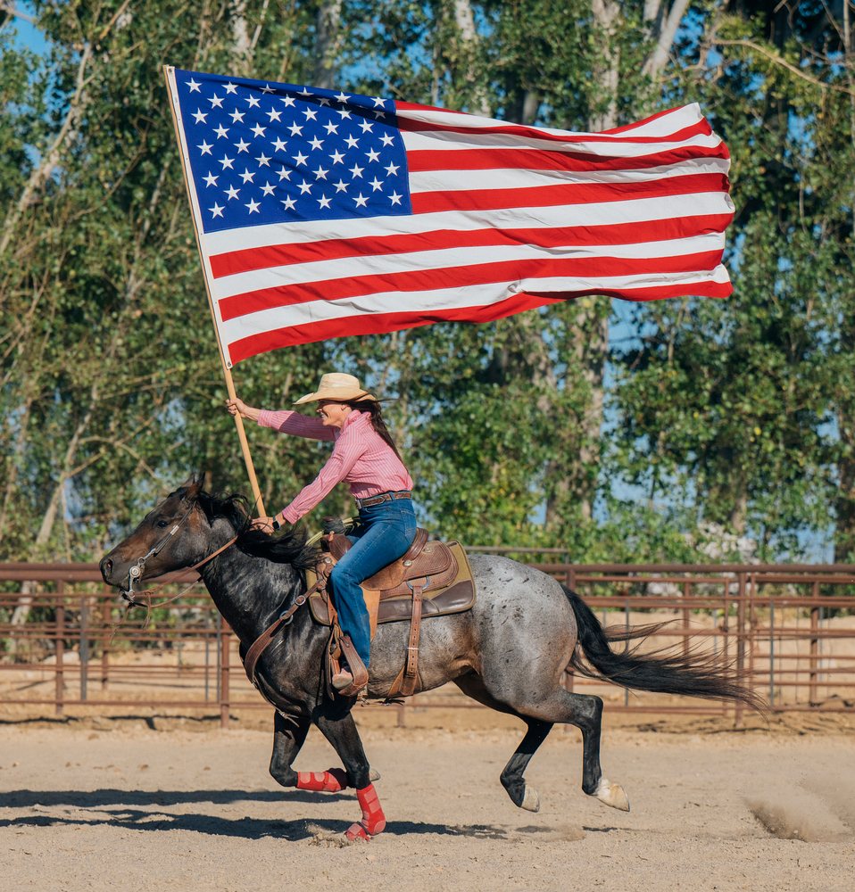 Woman wearing a cowboy hat rides a horse in a fenced outdoor arena holding a large American flag on a sunny day, Southwest USA.