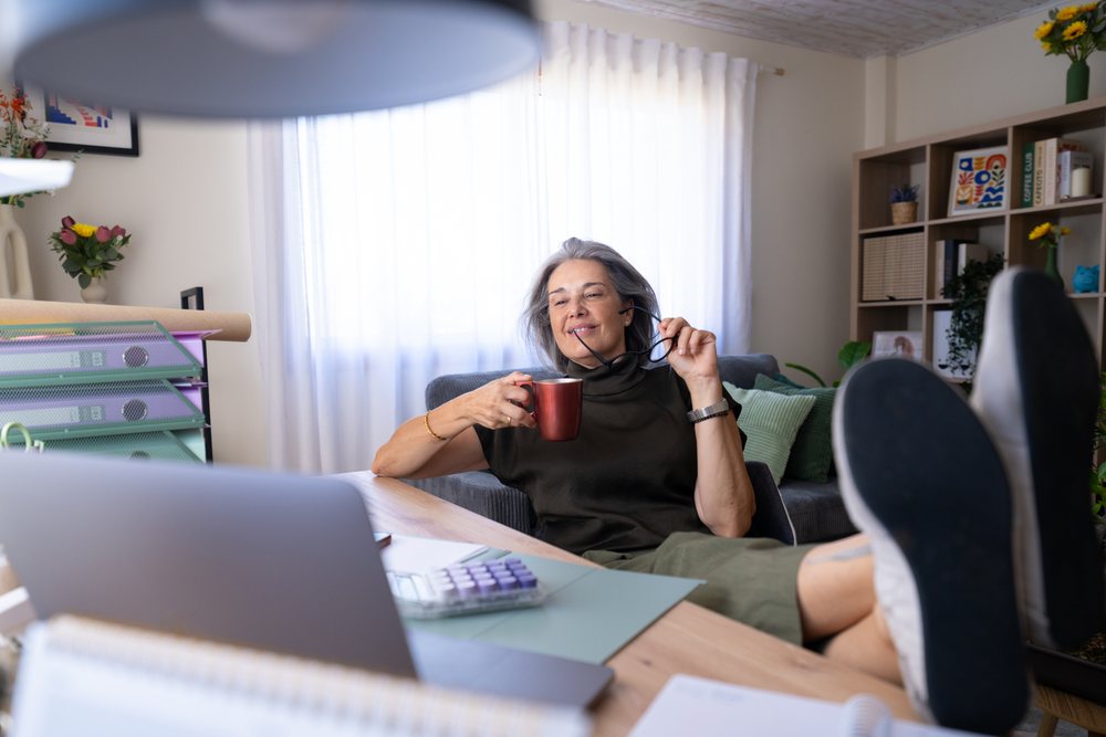 Woman sitting in a home office holding a coffee cup and glasses, wearing a headset with laptop on desk, Spain.