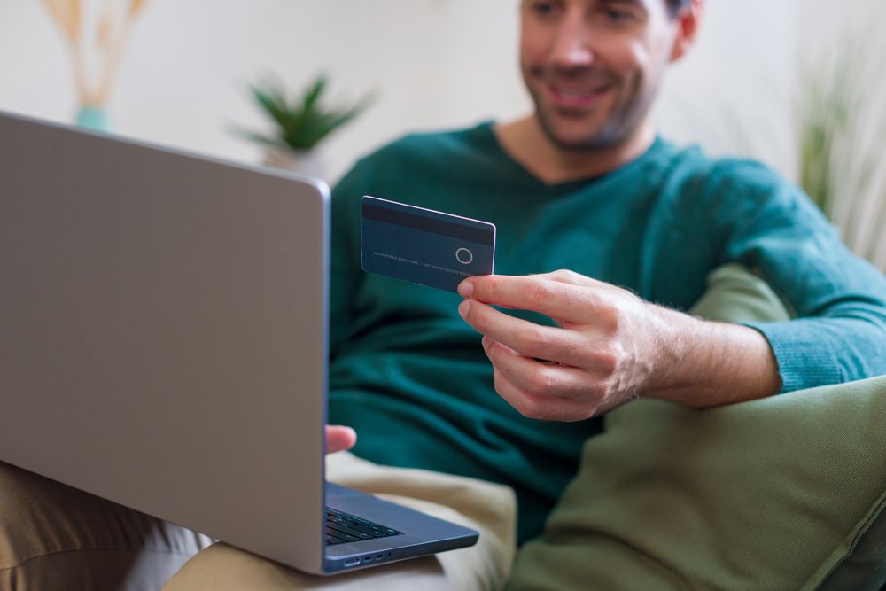Man wearing green sweater holding credit card and using laptop while sitting on sofa at home, technology, young adult.