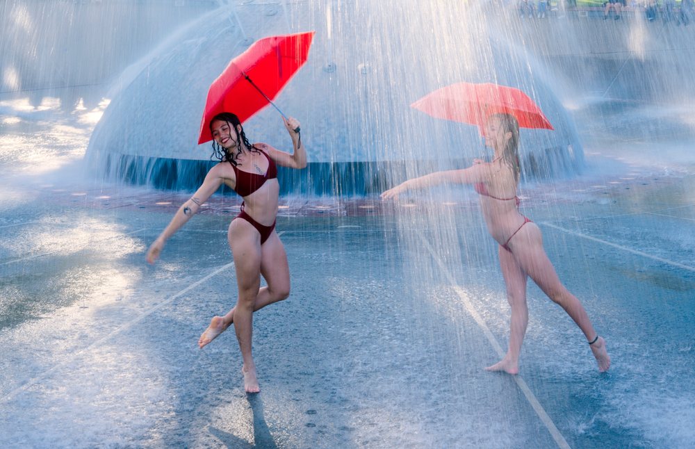 Two women dancing with red umbrellas under a water fountain spray in an outdoor urban park, partner, acrobatics, yoga, USA.
