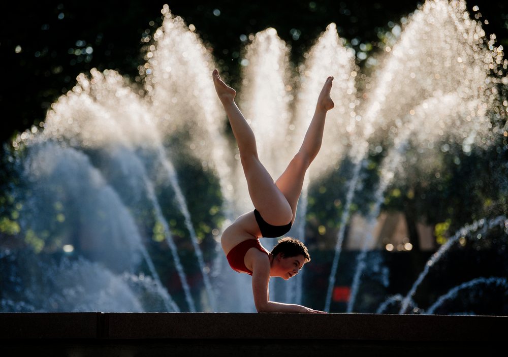 Person performing a handstand yoga pose outdoors on a stone platform with water fountains spraying, USA.