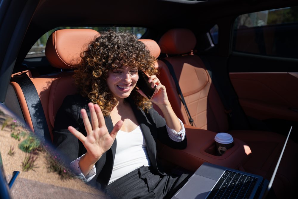 Businesswoman smiling and talking on phone while working on laptop in car back seat with brown leather interior, remote work, Spain.