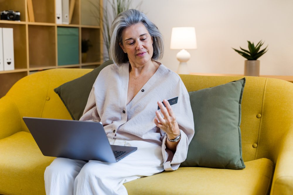 Woman with grey hair sits on a yellow sofa using a laptop and holding a credit card in a modern indoor living room, Spain.