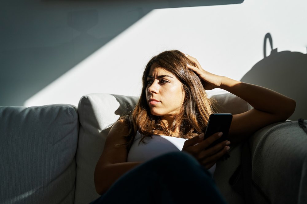 Young white woman holding mobile device sitting thoughtfully on couch