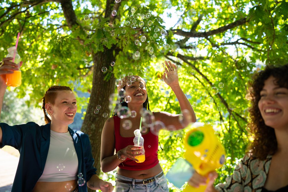 Three women outdoors under green trees enjoying bubbles and drinking orange juice in sunlight, Spain.