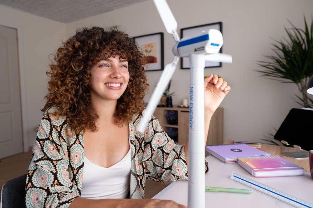 Girl with curly hair smiles as she examines a small model wind turbine on a desk with notebooks and a pen in a modern home interior, Spain.
