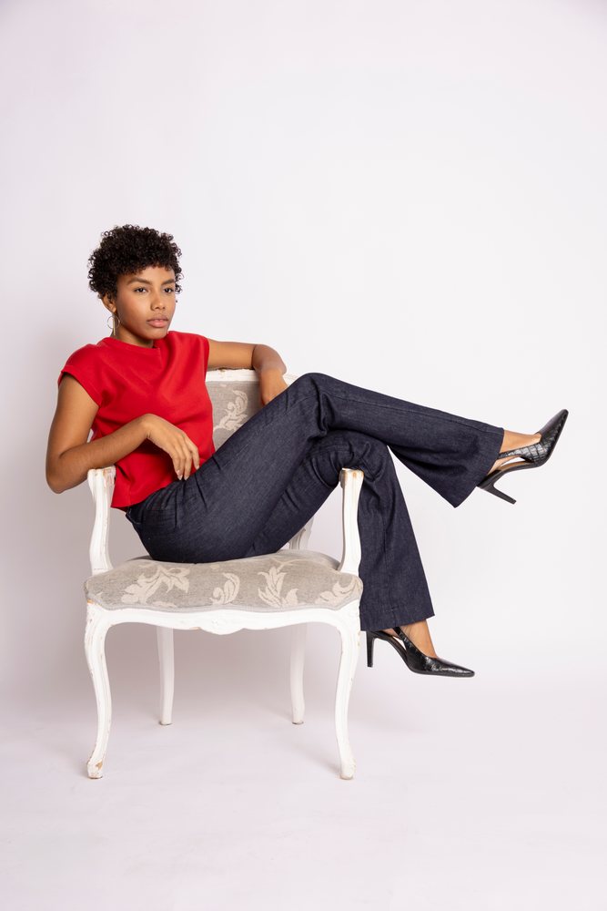 Woman with curly afro hair wearing a red top and black pants sits on a vintage chair in a studio, fashion, clothes, young culture.