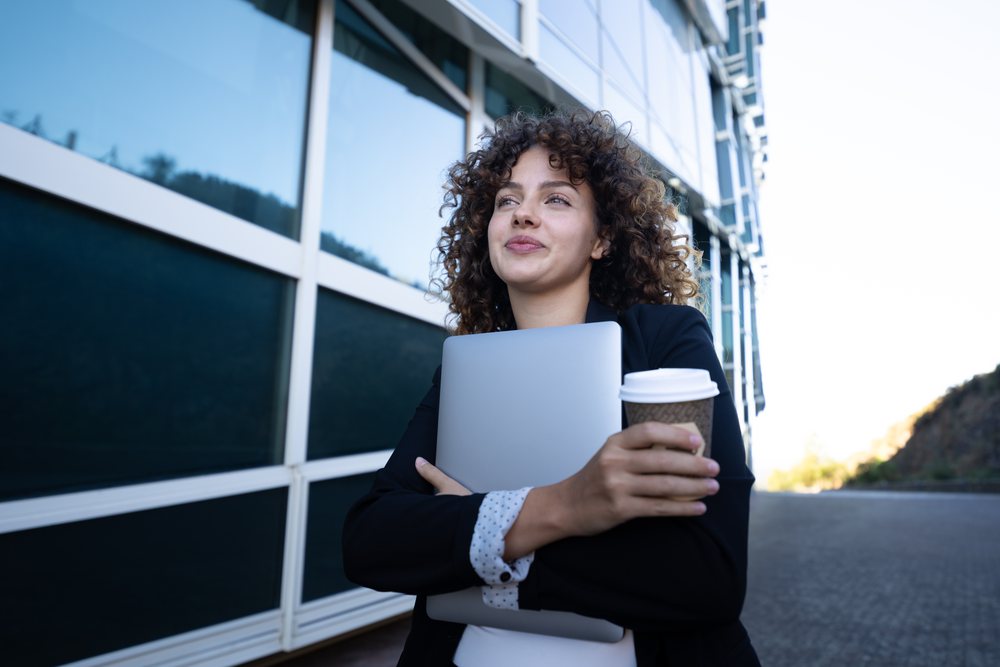 Businesswoman holding a laptop and a coffee cup outside a modern office building, business trip, Spain, Santiago De Compostela.