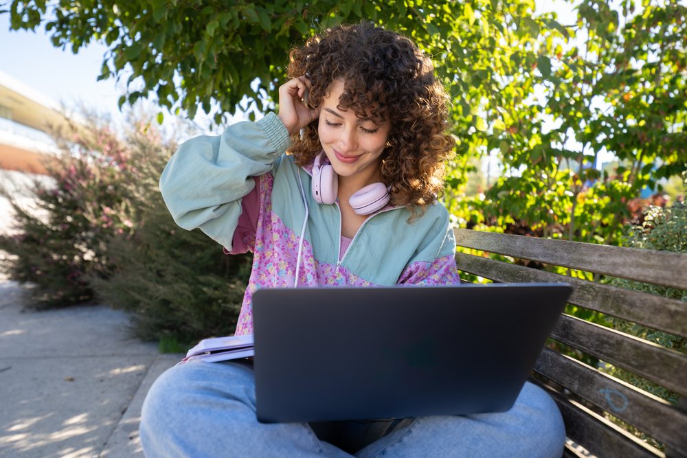 Woman sitting on a bench outdoors using a laptop with headphones around her neck on a sunny day in a park, Spain.
