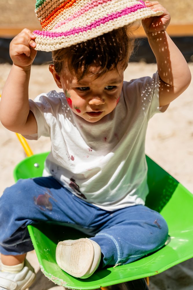 Child with strawberry face paint sits in a green wheelbarrow outdoors in sand under sunlight, kids, Spain.