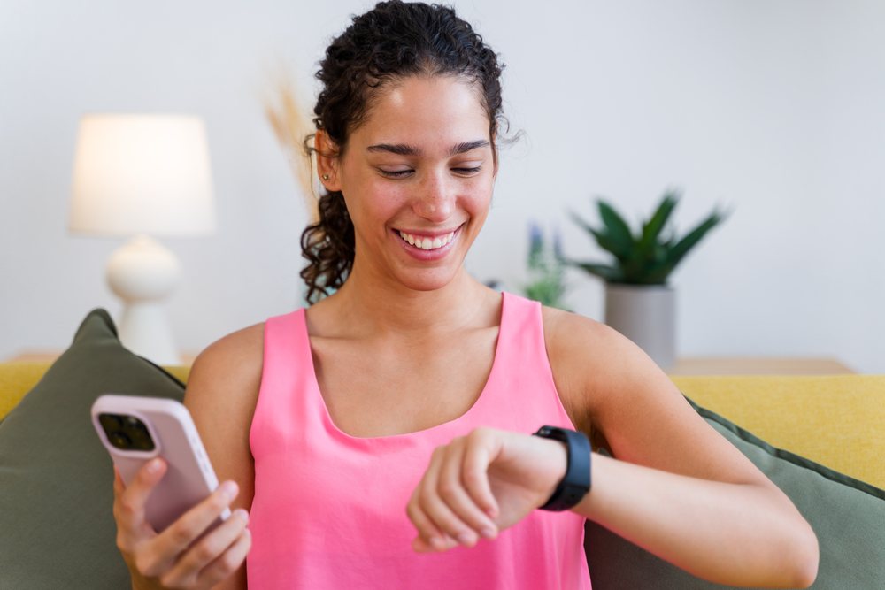 A smiling woman in a pink tank top checks her watch while holding a smartphone seated on a yellow sofa in a living room, Spain.