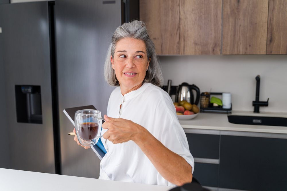 Middle-aged woman holding a coffee mug and tablet while seated in a modern kitchen, Spain.