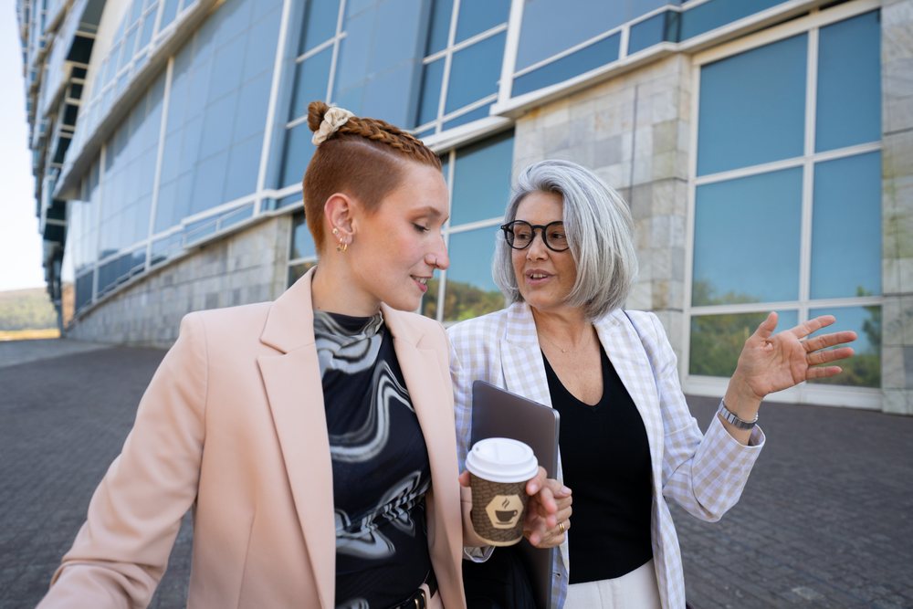 Two businesswomen walk and talk outside a modern office building, one holding a coffee cup and the other a tablet computer, Spain.