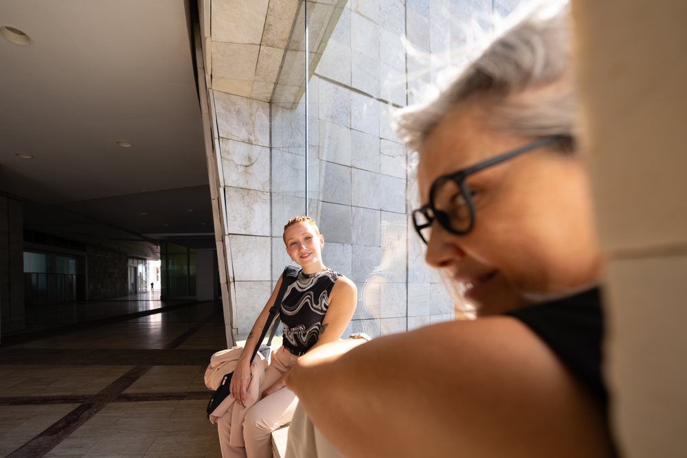 Two businesswomen communicate and smile beside a large modern glass window in an urban architectural walkway, Spain.