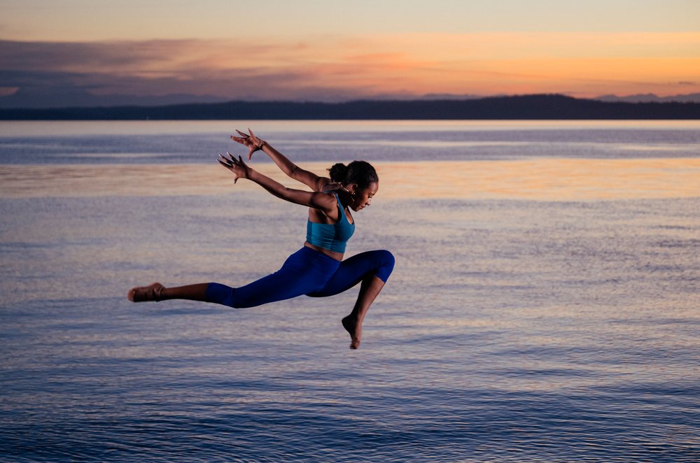 Woman performing a yoga acrobatic leap over calm ocean water at sunset, USA.