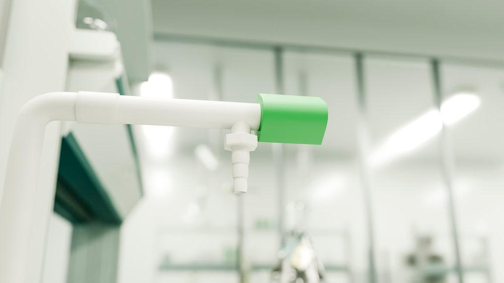 White valve with green handle attached inline on a laboratory pipe system with metal fittings and glass background elements.