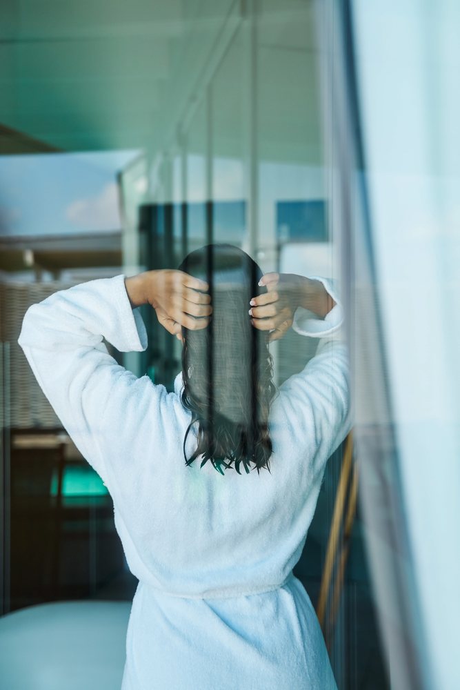 A woman in a white bathrobe stands indoors with her hands in her wet hair, seen through a glass door in the, Maldives.