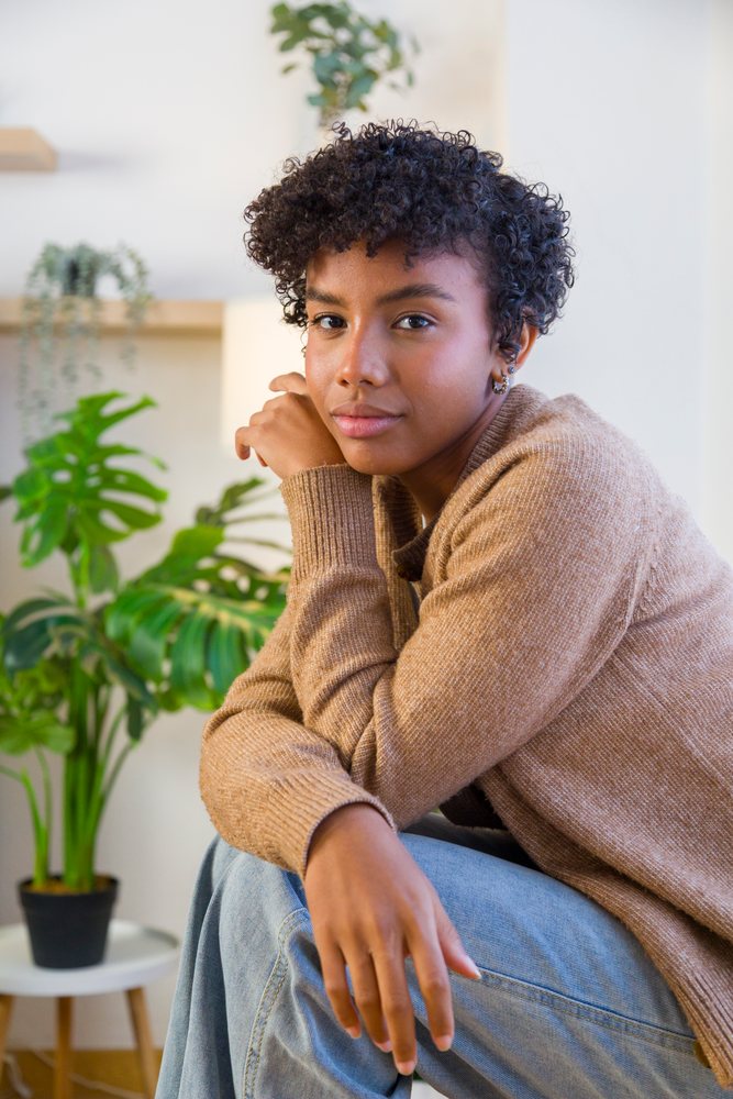 Young woman wearing a brown sweater and blue jeans sits indoors beside a Monstera plant in a cozy home setting.