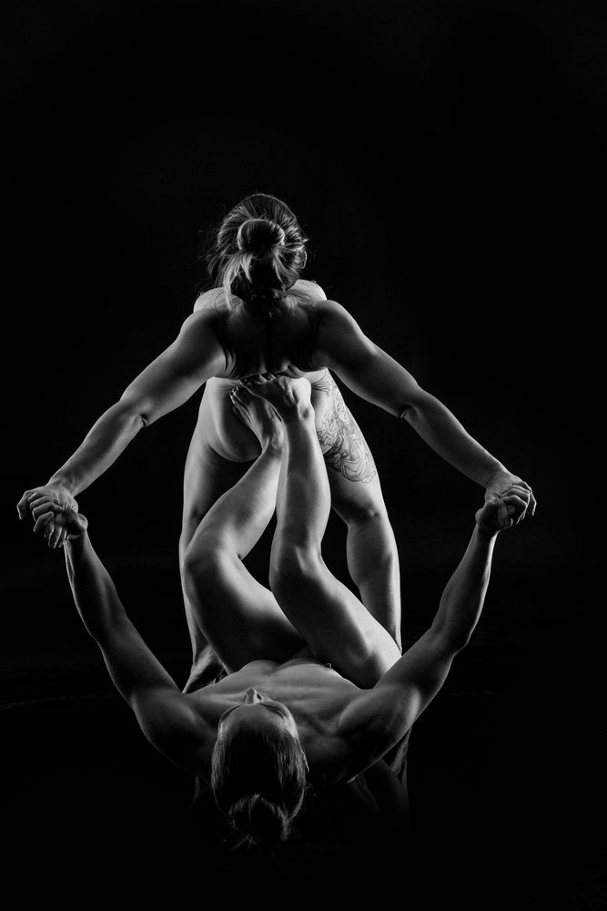 Two athletic individuals performing a partner acrobatics yoga pose balancing in a studio with a black background, body positivity.