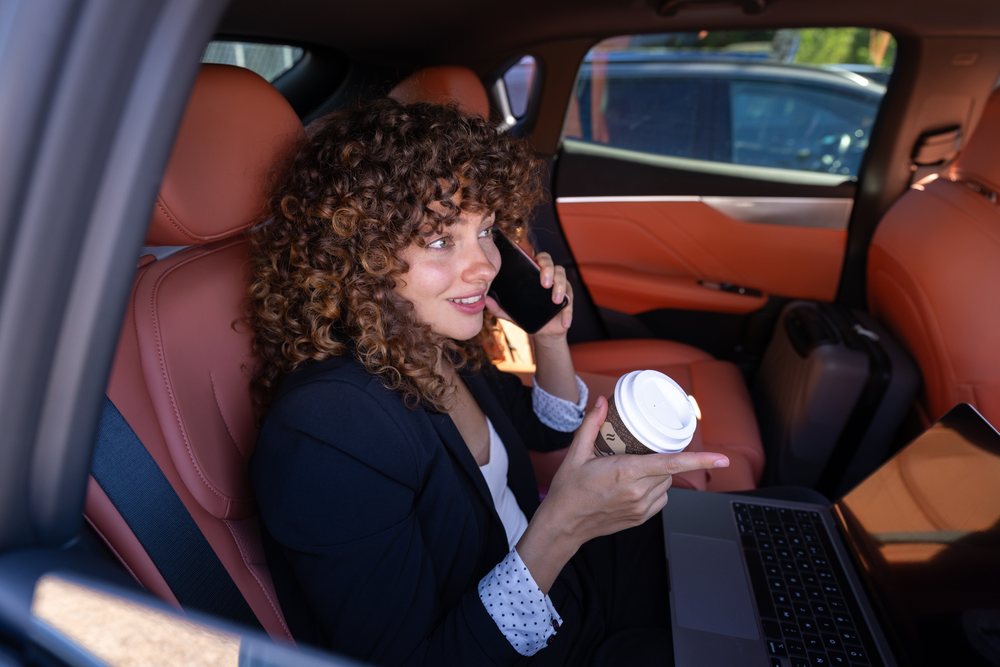 Businesswoman with curly hair works on laptop and talks on phone while holding coffee inside a car in Spain during daylight, remote work, businesswoman, car.