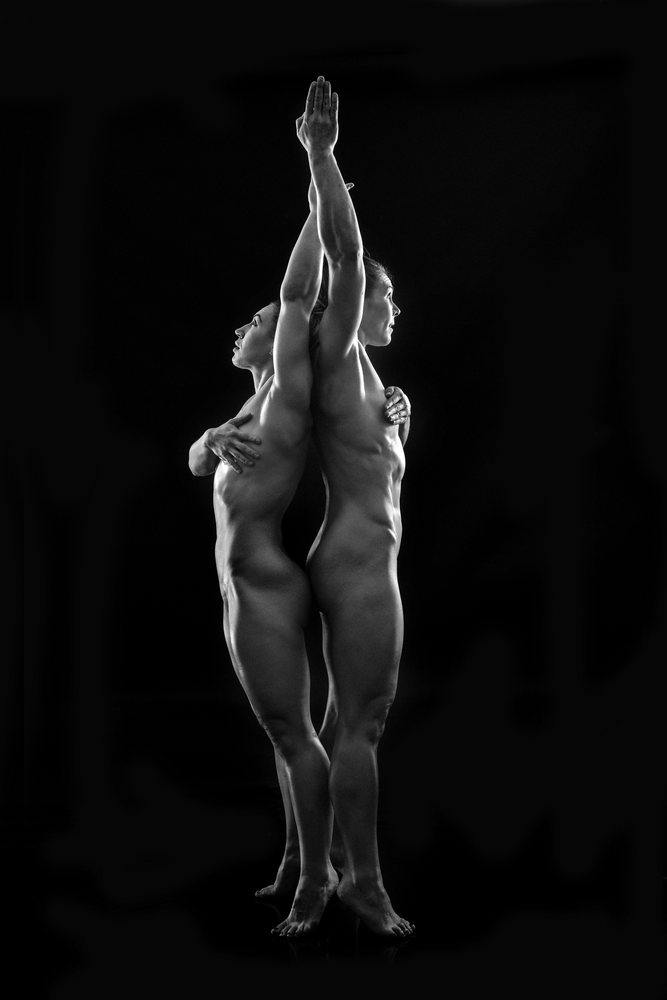 Two nude women performing a balanced standing back-to-back partner acrobatics yoga pose against a black background, body positivity.