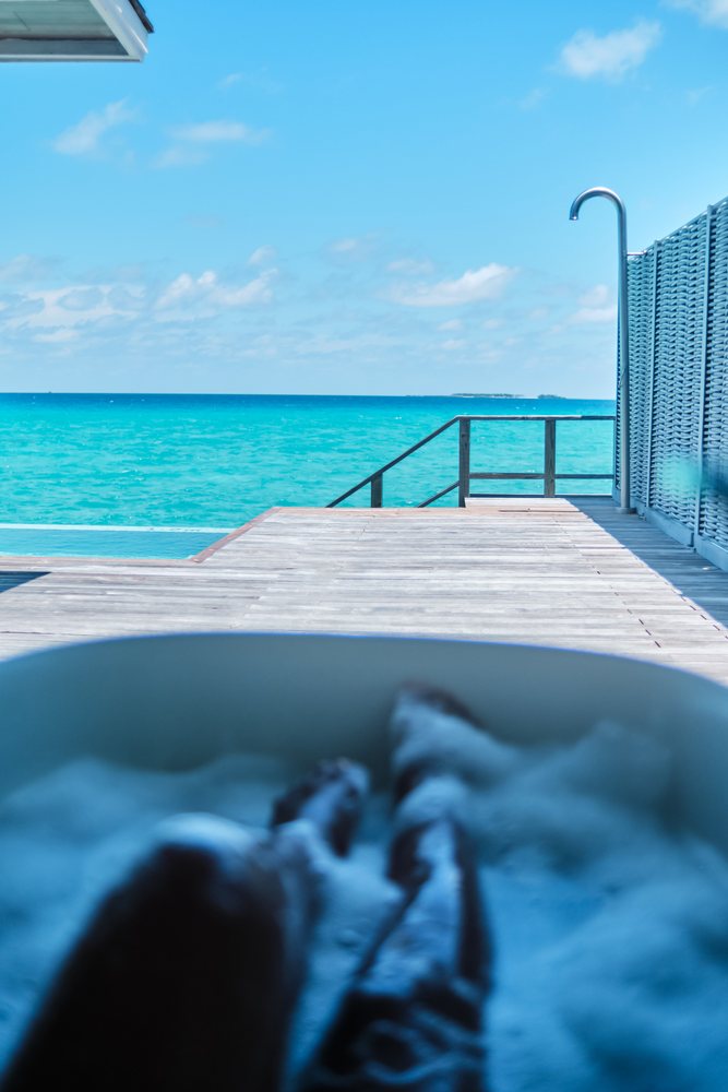 A person relaxes in an outdoor bathtub with bubbling water overlooking the turquoise ocean and clear blue sky in the, Maldives.