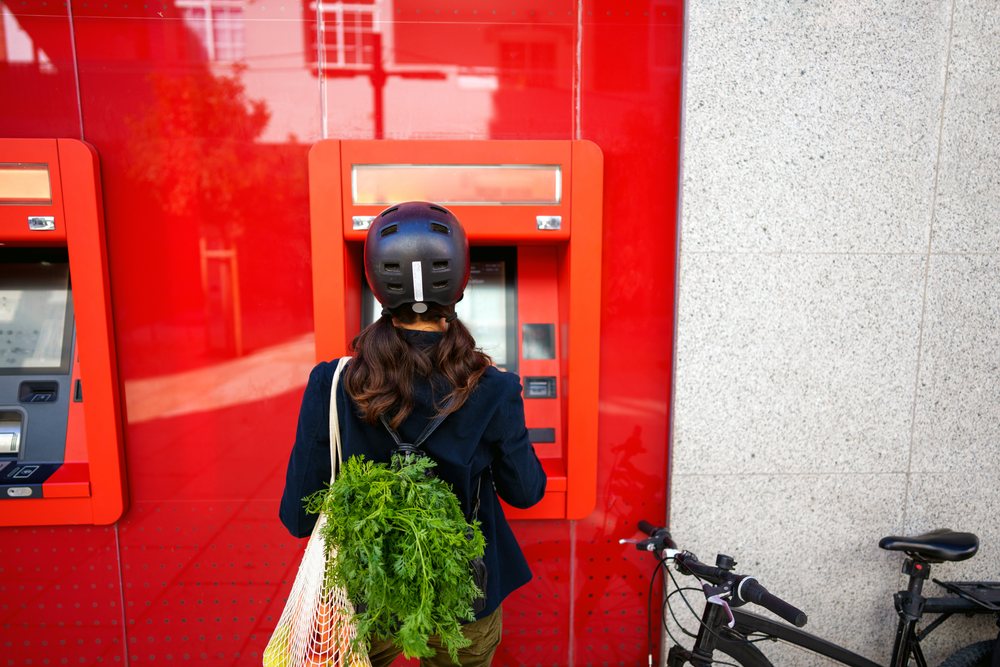 Person wearing helmet uses ATM outdoors with bicycle parked beside and green leafy vegetables in shopping bag, Málaga.