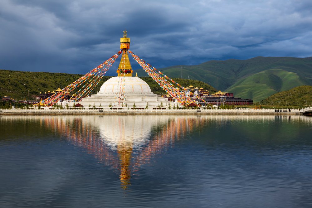 Large white stupa with golden pinnacle and colorful prayer flags reflects in a lake with mountains in the background at Zhuqing Wujian Zen Forest Monastery, Sichuan Province, People's Republic of China.