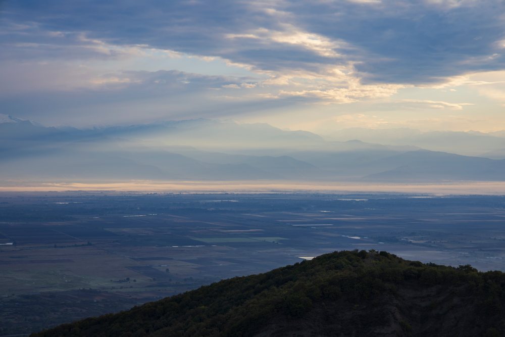 Sunrise over Alazani Valley, showing misty mountains, rural fields, and light rays through fog, Sighnaghi, Georgia.