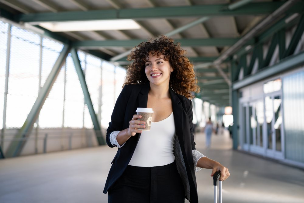 Smiling woman with curly hair walks through a corridor holding a coffee cup and pulling luggage, Spain.