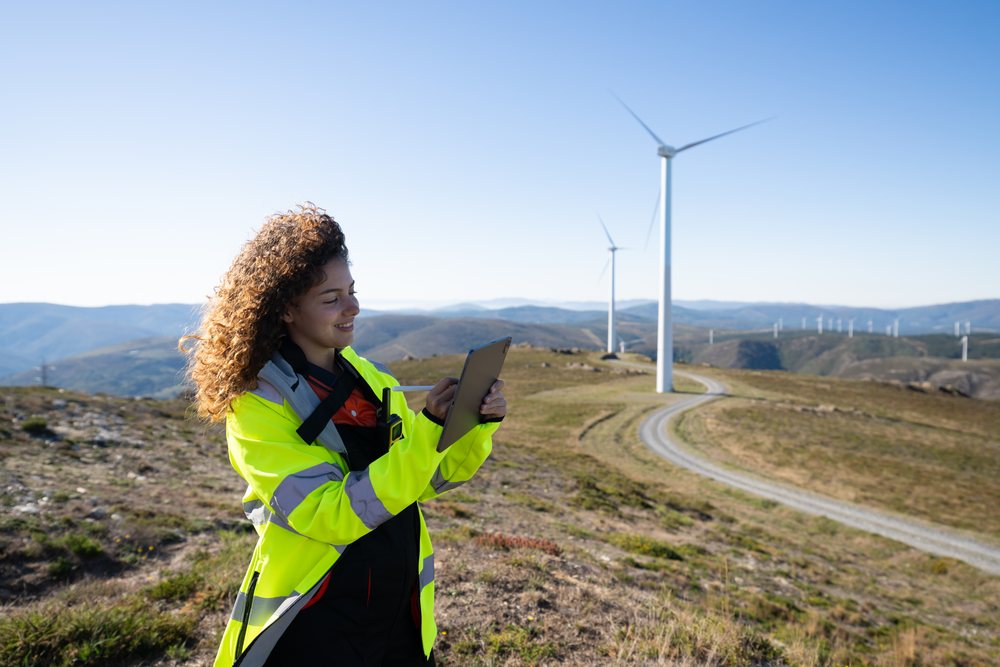 Woman wearing a high visibility jacket uses a tablet with digital pen at a wind farm on a hill during daytime, sustainability, renewable energy, Galicia, Spain.