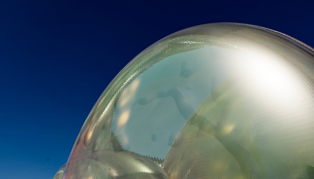 Glass sculpture with a distorted reflection of a figure on a curved shiny surface against a clear blue sky.
