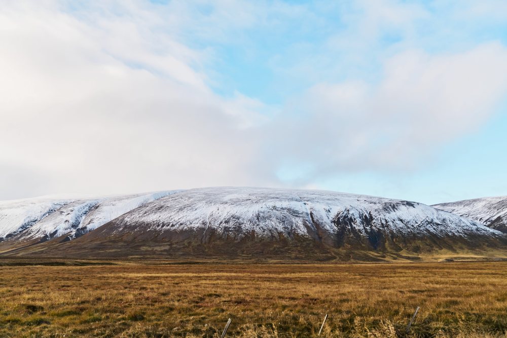 Snow-capped mountain rises above a vast grassland field under a blue sky, Iceland.